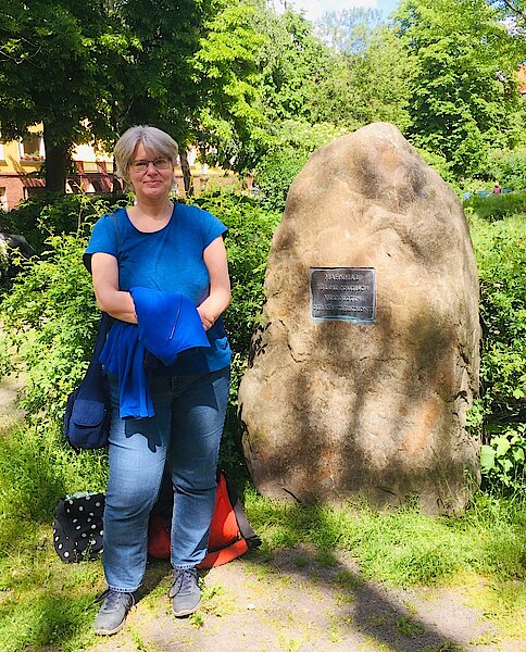 Doris Herrmanns steht neben dem Gedenkstein mit der Plakette, die an die politischen Verfolgten der Künstlerkolonie gedenkt.